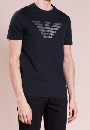 Man wearing black short-sleeve T-shirt with large silver stylized eagle logo and letters "GA" on chest, paired with black pants.