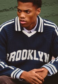 Námornícky modrý pletený svetr s nápisem "BROOKLYN" v bílé, s pruhovaným límcem a manžetami. Nositý přes světle modrou košili s límečkem.