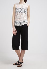 Top blanco sin mangas de encaje con patrón floral, combinado con culottes negros de pierna ancha y sandalias planas negras.