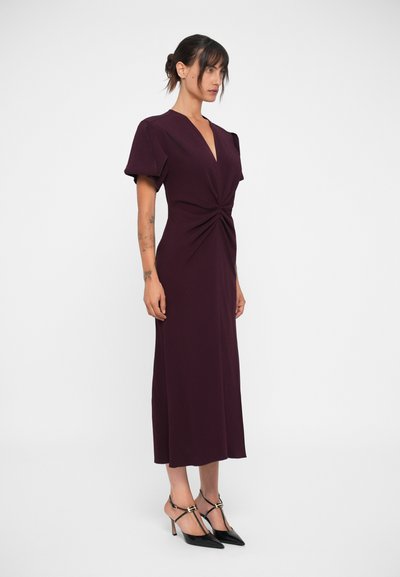 Burgundy midi dress with a deep V-neck and knot detail at the waist, featuring short puff sleeves and a sleek silhouette, paired with black heels.