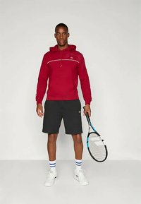 Κόκκινη hoodie με λευκή ρίγα, σε συνδυασμό με μαύρες βερμούδες. Κρατάει μία ρακέτα τένις, φορώντας άσπρα παπούτσια και μπλε ριγέ κάλτσες.