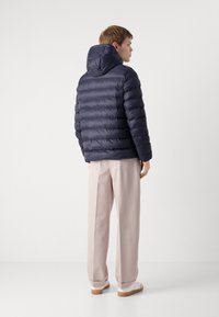 Giacca puffer navy con cappuccio, caratterizzata da un design trapuntato e una texture morbida. Indossata con pantaloni chiari e sneakers, vista di spalle.
