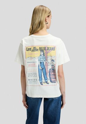 Kobieta z długimi blond włosami ma na sobie białą koszulkę z vintage'ową reklamą niebieskich jeansów Lee na plecach, zestawioną z niebieskimi jeansami.