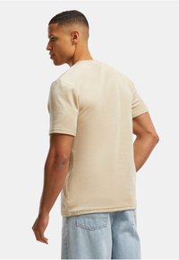 Camiseta de algodón beige con mangas cortas, cuello redondo y textura suave. El modelo está ligeramente girado, mostrando el diseño de la parte trasera.