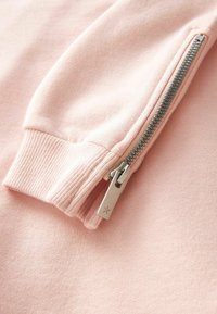 Svetle růžová mikina z látky se žebrovanými manžetami a stříbrným zipem na boku. Zip má jednoduchý vyrytý detail hvězdy.