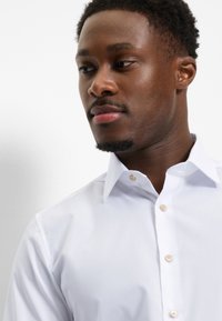 Young man with short curly hair wearing a white buttoned dress shirt, looking slightly to the side against a plain white background.