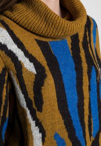 Sweter dziany z podwiniętym kołnierzem, w odcieniach brązu, niebieskiego, czarnego i białego, z wyrazistymi, abstrakcyjnymi wzorami i miękką teksturą.