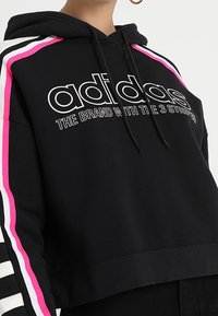 Černá zkrácená mikina s kapucí s bílým logem, zdobená růžovými a bílými pruhy na rukávech. Vyrobena z měkkého materiálu s nastavitelnými stahovacími šňůrkami.