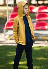 Żółta bluza z kapturem na zamek, noszona na granatowej koszulce, do tego ciemne spodnie typu cargo. W tle czerwone siedzenia na stadionie.