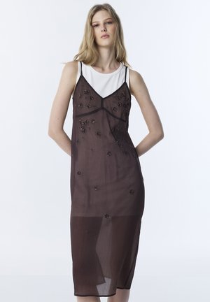 Vrouw draagt een doorschijnende donkerbruine slipjurk met bloemrijke kralenborduursels over een witte mouwloze top, staand tegen een effen lichte achtergrond.