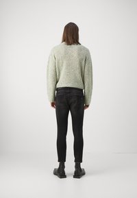 Persona con el cabello largo que lleva un suéter texturizado de color gris claro, jeans ajustados negros y botas negras hasta el tobillo, de pie de espaldas sobre un fondo sencillo.