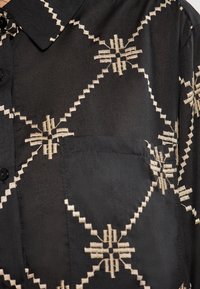 Camicia nera con colletto a punta, caratterizzata da un intricato ricamo beige in un motivo geometrico e una tasca sul lato sinistro.