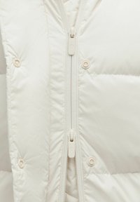 Pehme ja kohev kreemivärvi jakk kõrge kraega, täispikk tõmblukk ja nupud; omab siledat, läikivat tekstuuri ja õmmeldud detaile.