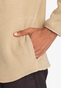 Pullover in pile beige con superficie morbida e texture; presenta una tasca laterale all'anca e orlo delle maniche a costine.