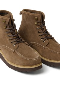 Stivaletti in suede marrone con punte rotonde, suole in gomma marrone scuro e occhielli in ottone per i lacci. Cuciture bianche accentuano il design.