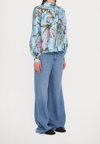 Persona con una blusa a maniche lunghe blu con fiori, jeans chiari a gamba larga e scarpe slip-on nere con pois bianchi, in piedi di lato.