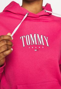 Sudadera con capucha de color rosa, hecha de algodón, que presenta un logo blanco de "TOMMY" con "JEANS" debajo, y cordones con puntas de metal.