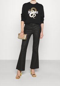 Kobieta w czarnych spodniach i czarnej bluzie z ze złotym wzorem węża, trzymająca małą złotą torebkę i nosząca złote sandały na wysokim obcasie.