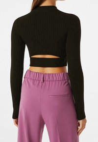 Kvinna i svart ribbat långärmad cropped topp med urtagning på ryggen och högmidjade lösa lila byxor, sedd bakifrån.