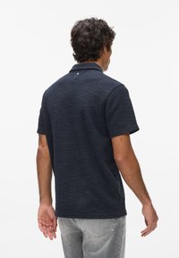 Granatowa koszulka polo z krótkim rękawem o teksturowanym splocie, z kołnierzykiem i małym logo z tyłu. Nosi ją z jasnoszarymi dżinsami.