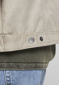 Svetle béžová semišová bunda se šedými patentkami, vrstvená přes tmavě zelený pletený svetr, v kombinaci se světle modrými džíny. Texturovaná tkanina, detailní pohled.