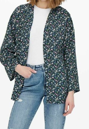 Woman wearing a dark floral open-front blazer over a white t-shirt and light blue high-waisted jeans with one hand in pocket.