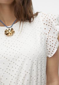 Blusa bianca in pizzo con maniche arricciate e un motivo testurizzato. Indossata con un pendente metallico dorato su una collana in corda blu.