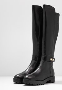 Bottes en cuir noir montant jusqu'au genou, avec une pointe arrondie, un accent doré sur la bride, et une semelle noire robuste. Texture lisse et design élégant.