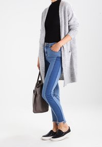 Ljust grå stickad cardigan över en svart polotröja, kombinerad med blå skinny jeans, svarta slip-on skor och en grå handväska.