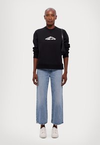 Black sweatshirt with white logo, blue cropped jeans with frayed hems, and white sneakers. Model carries a black shoulder bag.