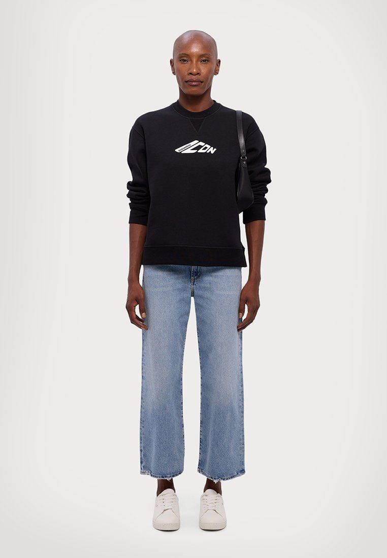 Sweatshirt noir avec logo blanc, jean bleu court avec ourlets effilochés, et baskets blanches. Le modèle porte un sac à bandoulière noir.