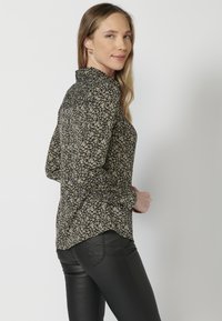 Blusa nera a maniche lunghe con un motivo floreale in giallo e beige. Presenta un colletto alto e un orlo curvo. Abbinata a pantaloni neri.