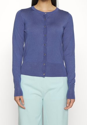 Femme portant un cardigan bleu cintré à manches longues boutonné, associé à un pantalon taille haute bleu clair, debout devant un fond uni.