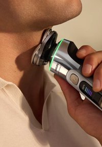 Elektrische scheerapparaat met een zilver- en zwarte ontwerp, voorzien van een groene LED-lamp, een meerbladige kop en een digitaal scherm voor de batterijstatus, tegen de huid gehouden.