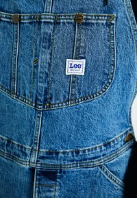 Blue denim overalls featuring a rectangular patch with the "Lee" logo, brass buttons, and prominent stitching details.