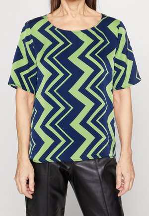 Mujer vestida con una blusa de manga corta azul marino y verde con un patrón de zigzag, usando pantalones de cuero negros, de pie contra un fondo liso.