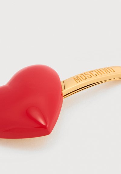 Accessoire rouge en forme de cœur attaché à une barre dorée gravée "MOSCHINO" sur fond blanc.