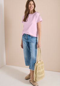 Femme souriante portant un haut rose clair, un jean bleu raccourci, des chaussures beige à enfiler, et tenant un sac cabas beige tissé.