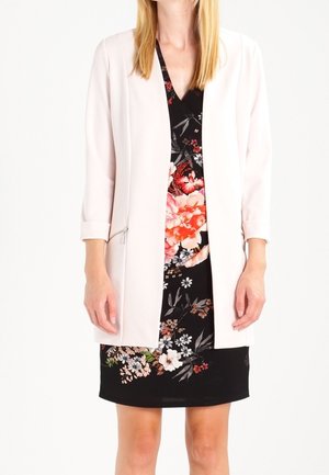 Femme portant une robe noire à fleurs rouges, roses et blanches, associée à un blazer rose clair ouvert devant avec des manches trois-quarts.