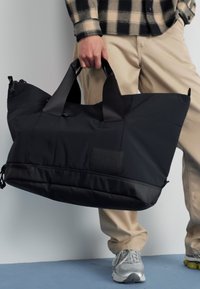 Black duffel bag with padded sides, top handles, and a logo patch. Made of durable fabric, features a zip closure and drawcord details.