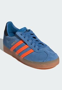 Zili zamšveida sporta apavi ar oranžām svītrām, melnu papēdi un gumijas zoli. Aprīkoti ar šņorēm, noapaļotu purngalu un "Gazelle" zīmola logotipu sānos.