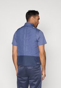 Veste légère bleue avec une partie supérieure matelassée et une partie inférieure unie. Coupe ajustée avec un col montant et un logo discret sur l'ourlet.