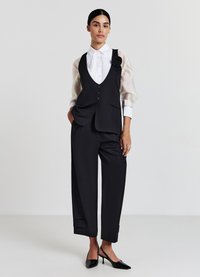 Schwarze maßgeschneiderte Weste mit Knöpfen, kombiniert mit weitem schwarzen Hosen. Weißes Hemd mit Kragen, Falten und durchsichtigen Puffärmeln. Schwarze High Heels.