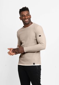 Beige Langarmshirt mit strukturiertem Rippdesign, Rundhalsausschnitt und einem schwarzen Akzent am linken Ärmel. Kombiniert mit schwarzen Hosen.