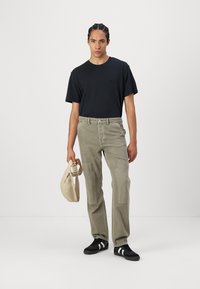 Svart t-shirt, ljus olivgrön cargobyxor med fickor fram, strukturerade knädetaljer och svarta sportskor, håller i en beige väska.