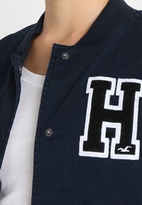 Námorně modrá bomber bunda s bílým a černým vyšitým písmenem "H" na hrudi, strukturovaná tkanina a zapínání na cvoky.