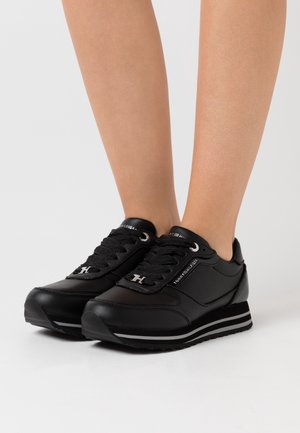 Jambes portant des baskets noires Tommy Hilfiger avec des accents argentés et le logo sur le côté et la languette, debout sur une surface blanche.