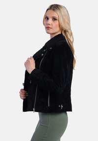 Žena stojí bokem, na sobě má černou semišovou bundu s huňatým límcem, kterou si rukou drží zavřenou, a je umístěna na jednobarevném pozadí.