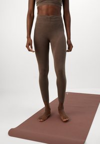Person standing barefoot on brown yoga mat, wearing brown high-waisted leggings and a matching crop top, against a plain light background.