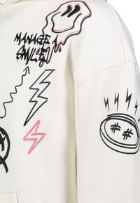 Crème-kleurige hoodie met verschillende zwarte en roze grafische ontwerpen, waaronder bliksemschichten, een geest en tekst die zegt "BEHEER EEN GLIMLACH."